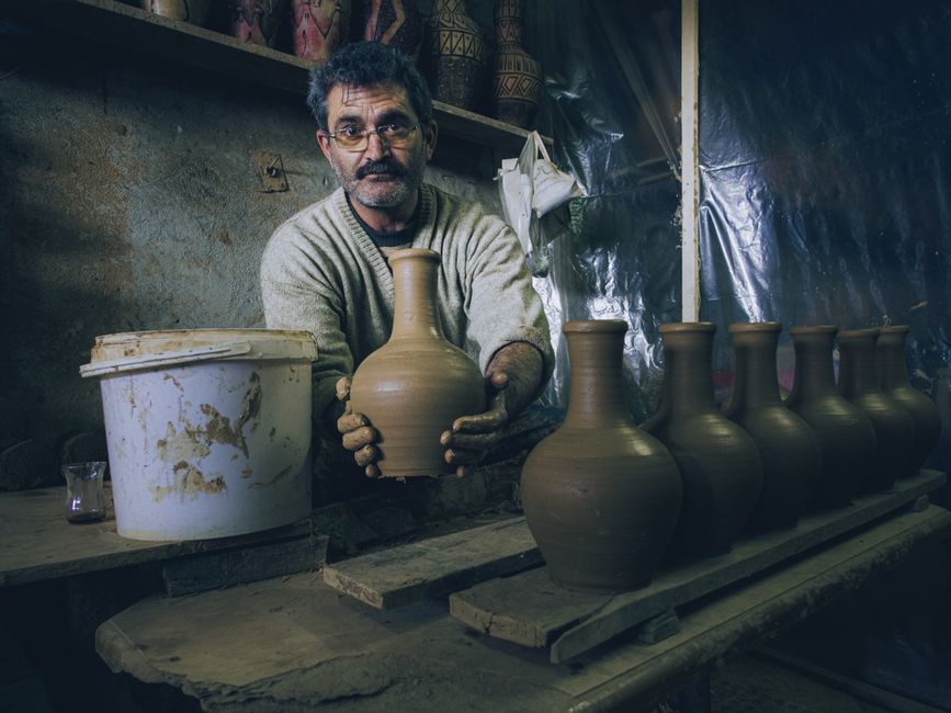 A craftsman in a workshop carefully shapes a clay vase by hand, surrounded by finished pottery.